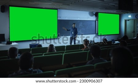 Young Male Teacher Giving a Lecture, Showing Slides on a Green Screen Mock Up Display to a Diverse Multiethnic Group of Female and Male Students in Dark College Auditorium