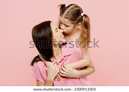 Happy lovely woman wear casual clothes with child kid girl 6-7 years old. Daughter stand behind mother, kiss nose, hug and cuddle isolated on plain pastel pink background. Family parent day concept