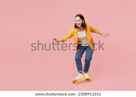 Full body young happy fun cool woman of Asian ethnicity wear yellow shirt white t-shirt riding skateboard pennyboard isolated on plain pastel light pink background studio portrait. Lifestyle concept