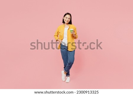Full body fun young woman of Asian ethnicity wear yellow shirt white t-shirt hold takeaway delivery craft paper brown cup coffee to go isolated on plain pastel light pink background studio portrait Royalty-Free Stock Photo #2300913325