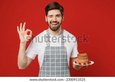 Young smiling happy fun male housewife housekeeper chef cook baker man wearing grey apron hold in hand plate with pancakes show ok gesture isolated on plain red background studio. Cooking food concept Royalty-Free Stock Photo #2300913525