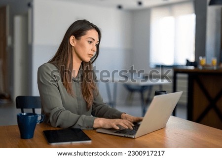 Serious dedicated businesswoman using laptop sitting at the table in a home office, looking at device screen, communicating online, writing emails, distantly working or studying on computer at home. Royalty-Free Stock Photo #2300917217