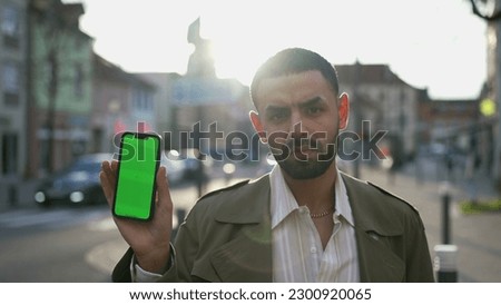 A serious young Arab Moroccan man holding cellphone with chroma greenscreen standing outside in city street. Blank smartphone device screen