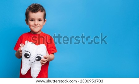 A smiling boy with healthy teeth holds a plush tooth in his hands on a blue isolated background. Oral hygiene. Pediatric dentistry. Prevention of caries. A place for your text. Royalty-Free Stock Photo #2300953703