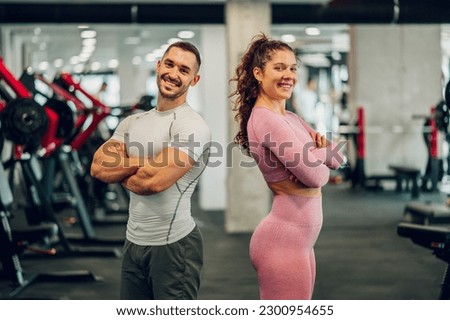 A happy gym friends are standing confidently with arms crossed back to back and smiling at the camera. A muscular couple is standing with their arms crossed in a sports center and making eye contact.
