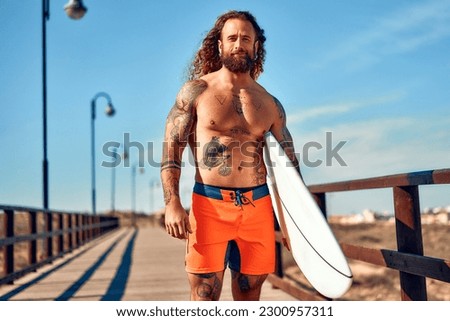 Caucasian beefy bearded man with tattoos in swimming trunks holding a surfboard walks along a wooden bridge to the sea. Sports and active recreation. Royalty-Free Stock Photo #2300957311