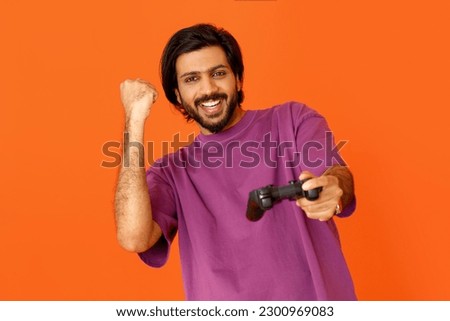 Addiction, gaming disorder concepy. Happy cheerful young indian guy in purple t-shirt playing video game, holding joystick console and gesturing, orange studio background, copy space. Gaming concept
