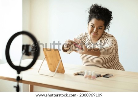 Pretty smiling curly hispanic overweight young lady makeup artist recording video for her beauty blog on smartphone, woman sitting at desk, testing brand new beauty products, showing lip gloss