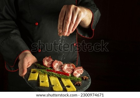 Professional chef sprinkles salt on a sliced steak with ham and cheese on a serving plate. The concept of serving dishes to order by a waiter with space for a menu on a dark background