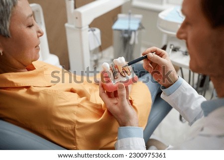 Closeup of male dentist holding tooth model while explaining dental implant surgery to patient in clinic, copy space Royalty-Free Stock Photo #2300975361