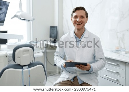 Portrait of smiling male dentist looking at camera in dental clinic interior and holding clipboard, copy space Royalty-Free Stock Photo #2300975471