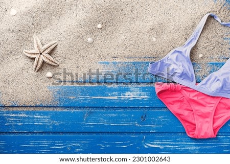 Flat lay shot of pink and purple two piece swimsuit. Sand and starfish on blue weathered wooden beach platform. 