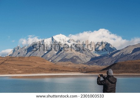 A man tourist in black jacket and a hat taking a picture of the view of Laguna Amarga, mountains in the national park Chilean Patagonia Torres de Paine