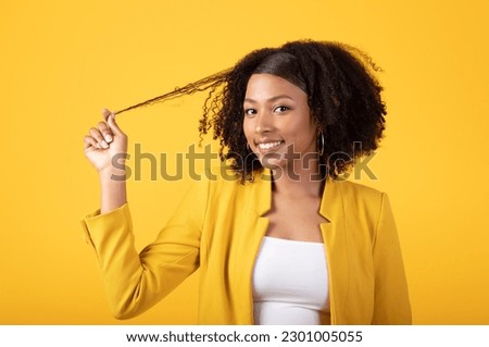 Portrait of happy african american lady touching hair and flirting, looking at camera and smiling isolated on yellow studio background. Human emotions, ad and offer