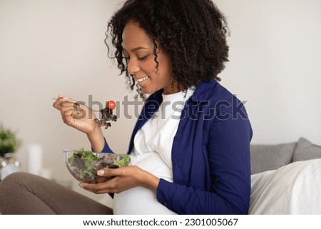 Smiling millennial african american lady with big belly eating salad in room interior, close up. Health care, proper nutrition, vegan and expectation of child, motherhood and family Royalty-Free Stock Photo #2301005067