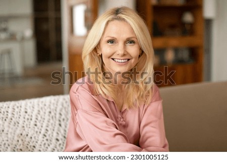 Portrait Of Blonde European Mature Woman Posing Smiling To Camera Sitting On Sofa At Home. Lady Resting On Cozy Couch In Modern Living Room Indoor. Ageless Beauty And Elegance Concept Royalty-Free Stock Photo #2301005125