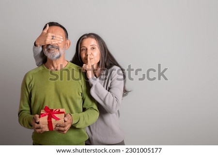 Glad caucasian elderly lady closes eyes to husband, make shh sign, gives gift, congratulations on holiday on gray studio background. Birthday greeting, anniversary, surprise, relationship and love