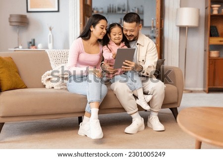 Online Leisure For Family. Happy Japanese Parents And Child Daughter Using Digital Tablet Browsing Internet And Watching Cartoons Online Sitting On Sofa At Home. Weekend Together Concept