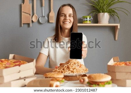 Delicious meal. Online ordering, Fast delivery. Smiling woman wearing white t- shirt siting at table with junk food in kitchen showing smartphone with empty display, copy space for advertisement.