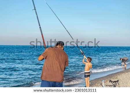 A young boy in swimming shorts and an unrecognizable man hold fishing rods on the pebble beach on sunny a summer day with intense blue water and clear sky.  A dressed boy washes his hands by the sea.