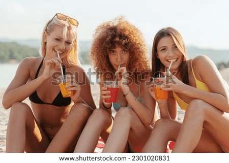 Three young women relaxing and enjoying on the beach, drinking juice and cheering. 