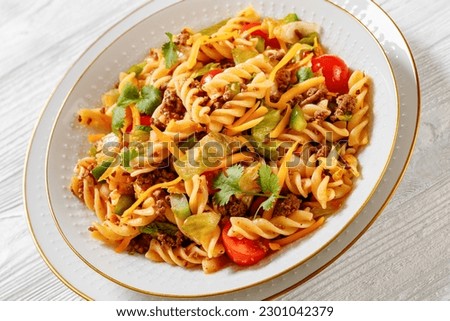 Nacho pasta salad with ground beef, tomatoes, green peppers, iceberg lettuce and taco seasoning in white bowl on white wood table, close-up