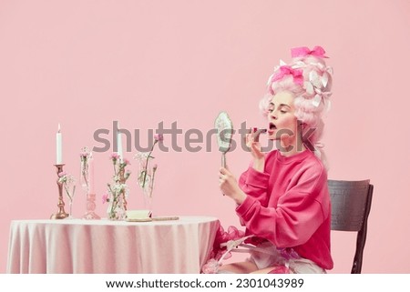 Beauty salon. Portrait with pretty princess, queen wearing big wig holding hand mirror and using lipstick on pink background. Concept of comparison of eras, modernity and renaissance, beauty, history Royalty-Free Stock Photo #2301043989