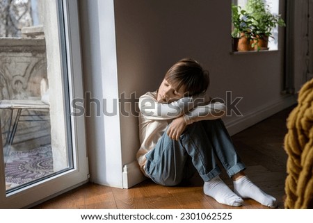 Tired sleepy teen boy sitting on floor embracing knees at home near window. Lost broken frustrated teenager having life problems troubles. Teenage crisis, abuse, domestic violence, depression concept. Royalty-Free Stock Photo #2301062525