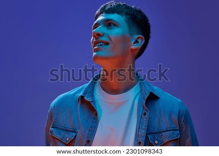 Portrait of young handsome guy in jeans shirt and white t-shirt design with smile, looking away against gradient purple background in neon light. Concept of human emotions, lifestyle, youth