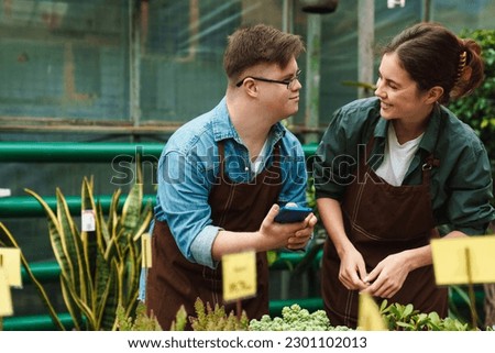 Young man with down syndrome and young woman florist wearing aprons smiling while working together in greenhouse Royalty-Free Stock Photo #2301102013