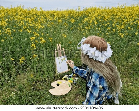 attractive young woman sitting in oilseed rape field. girl painting nature scenery on canvas. agriculture wallpaper.  lady wearing plaid shirt and flower wreath on head.