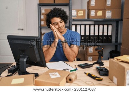 Hispanic man with curly hair working at small business ecommerce sleeping tired dreaming and posing with hands together while smiling with closed eyes. 