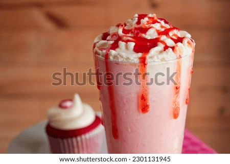 Close-up tall glass filled with a fresh strawberry milkshake, topped cream with additional strawberries jam for the perfect refreshing dairy dessert. Added red velvet cupcakes behind Royalty-Free Stock Photo #2301131945