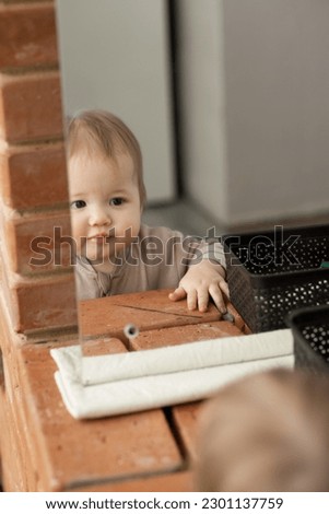 The kid looks at himself in the mirror standing on the red brick fireplace