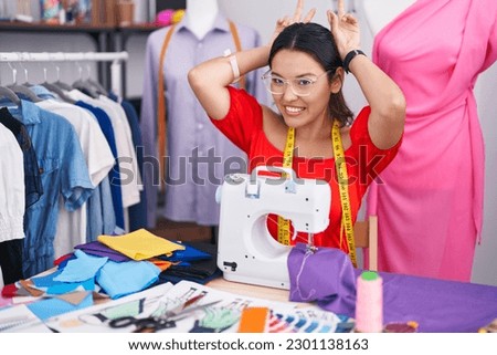 Hispanic young woman dressmaker designer using sewing machine posing funny and crazy with fingers on head as bunny ears, smiling cheerful 