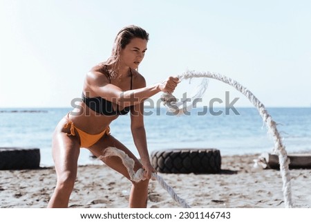 Young woman athlete exercising with battling rope during her workout at a beach gym.
