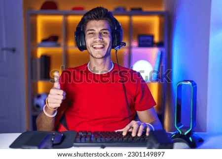 Young hispanic man playing video games doing happy thumbs up gesture with hand. approving expression looking at the camera showing success. 