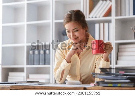 Pleasant young female employee drinking coffee and working with document at office desk.