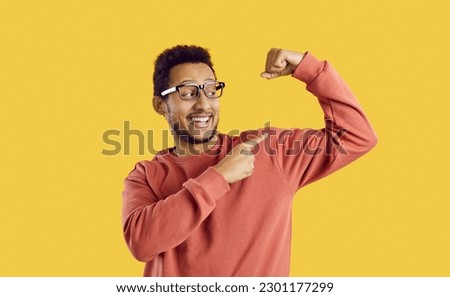 Portrait of funny young man showing off his biceps on his arm isolated on bright yellow background. Close up of smiling african american guy in glasses and sweater showing finger on his muscles. 