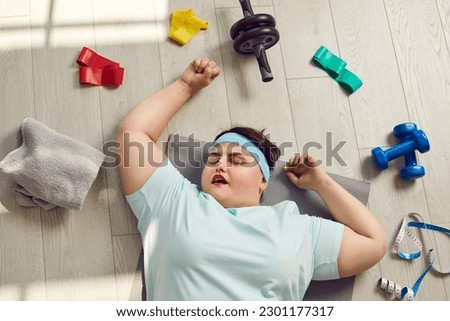 Tired lazy fat woman falls asleep during fitness workout. Funny overweight plump woman sleeping on floor among sports equipment. Top view high angle from above. Weight loss, lack of motivation concept Royalty-Free Stock Photo #2301177317