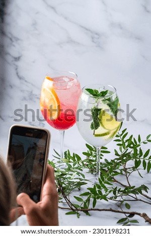 Fresh summer cocktails with fruits and ice cubes. Girl taking pictures on the phone cocktails Aperol Spritz with orange and Mojito with mint and lime. Vertical orientation.