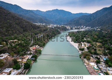 aerial drone shot of blue water of river ganga stretching into distance with himalayas with ram setu suspension bridge and temples on the banks of the river