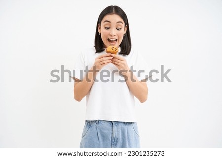 Image of funny asian woman, eating delicious cupcake, likes pastry, munching dessert with delight, white background.