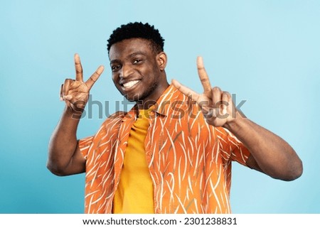 Portrait of handsome smiling African American man wearing orange shirt showing peace sign isolated on blue background. Young stylish hipster model looking at camera posing for pictures. Summer concept