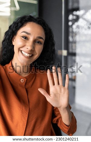 Vertical shot, young beautiful Hispanic business woman with curly hair talking on video call, waving at smartphone camera, using remote communication app. Royalty-Free Stock Photo #2301240211