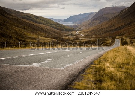 Ground level view of a winding road across a valley leading to a lake surrounded by misty mountains in the Scottish Highlands, United Kingdom