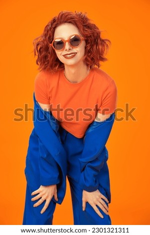 Bright colors. Portrait of a beautiful cheerful girl with foxy curly hair smiling laughing in bright blue and orange clothes and sunglasses. Studio orange background.