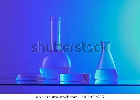 Front view of lab glassware containing colorless liquid and transparent podium displayed on blue background. Pedestal for cosmetic product presentation with laboratory concept. Royalty-Free Stock Photo #2301332685