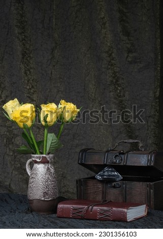 yellow roses in a vase against a background of dark fabric, next to an old wooden chest. leather-bound book and jewel. Place for text. Mystery, riddle.