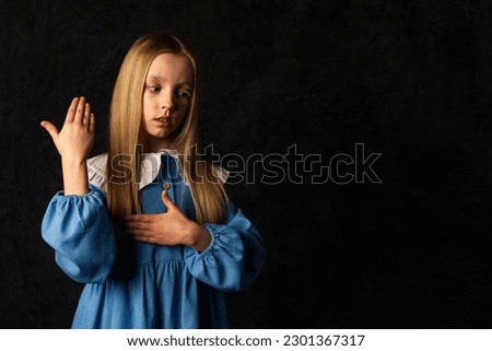 Portrait of cover little doll girl in blue dress gesturing hands at black background, looking away. Lovely child 9-10 year old in image isolated in dark. Fashion masquerade concept. Copy ad text space